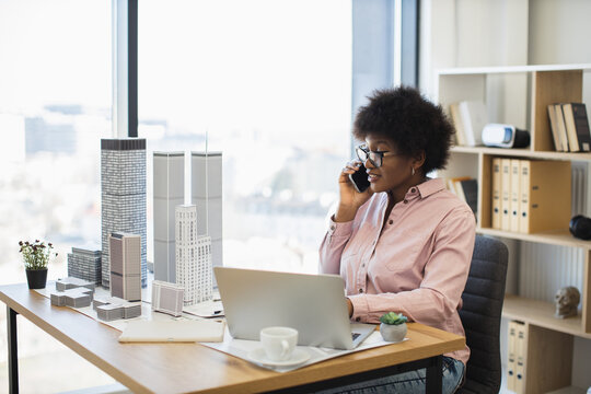 Confident African woman architect in office talking on smartphone about city project. Middle-aged professional surrounded by skyscraper models, working on laptop, creating future urban development.