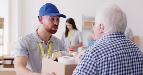 A volunteer guy gives a box of groceries to an elderly man, slowmotion. In the background, a group of volunteers in a warehouse collects groceries. Distribution of aid to those in need - Powered by Adobe