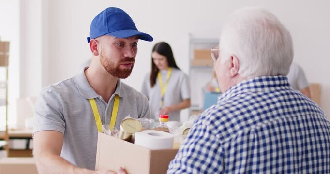 A volunteer guy gives a box of groceries to an elderly man, slowmotion. In the background, a group of volunteers in a warehouse collects groceries. Distribution of aid to those in need