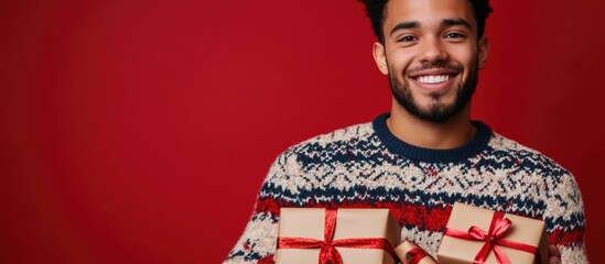 Fototapeta premium Young man in festive sweater holding gift boxes against vibrant red background celebrating holidays and joyful moments