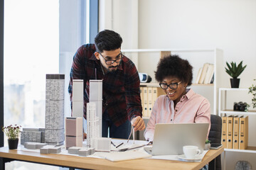 Indian man and African woman architects focused on innovative city project at desk. Collaborative teamwork in office setting with architectural models and laptop.