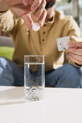 Close-up photo of young man dropping effervescent tablet into glass of water, with tablet dissolving sitting at table in living room. Concept of illness, healthy insurance, pharmaceutical, medicine.