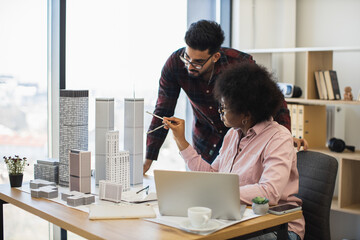 Indian man and African woman architects collaborate on model project for modern city. Diverse teamwork in bright office setting with creative discussion and urban planning focus.