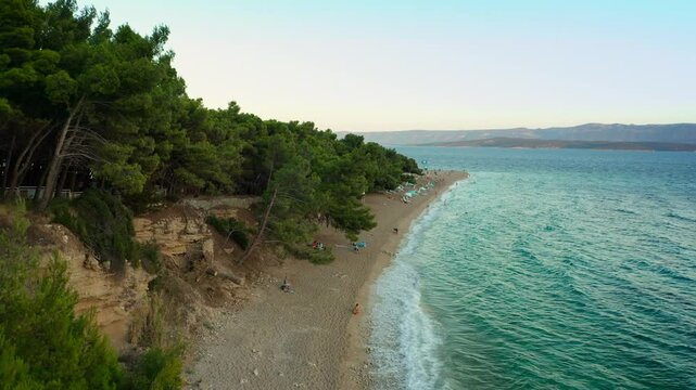 Zlatni Rat beach at sunset. People swim in the sea, swinging on the waves and watching the sunset, relaxing and sunbathing on the shore. Tourism and beach recreation on the Adriatic coast of Croatia.