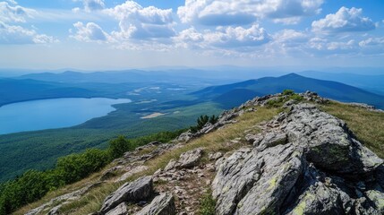 Scenic mountain landscape overlooking serene lake under a clear blue sky with fluffy clouds and lush green hills in the distance