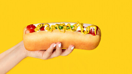 Cropped view of young girl holding yummy hot dog with mustard and ketchup on orange background