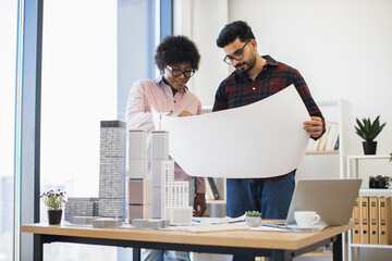 Indian man and African woman architects examining blueprints developing project for modern city. Office setting with architectural models, teamwork and collaboration emphasized.