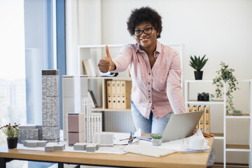 Confident African American female architect giving thumbs up in modern office setting with architectural models, showcasing success and creativity in urban design.