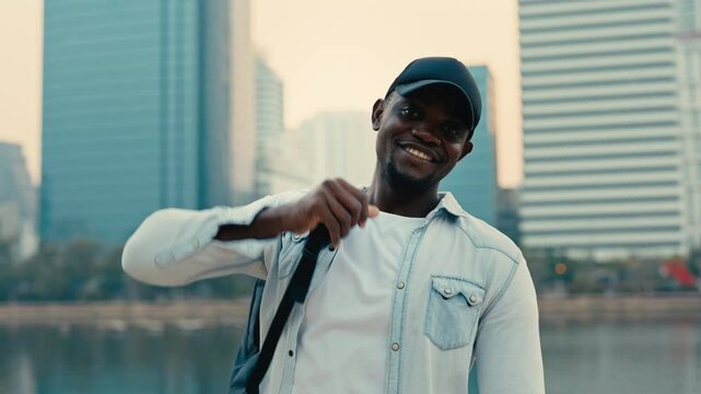 Portrait of happy man adjusting bag on shoulder against skyscrapers. African American guy with beard and baseball cap shows casual attire in city. Urban environment life