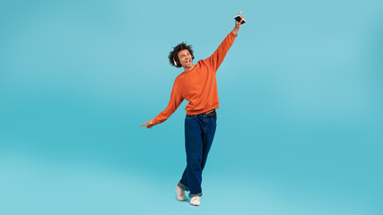 Obraz premium A young African American man with curly hair stands playfully against a vibrant light blue background. He raises one arm joyfully while holding a smartphone, expressing a carefree lifestyle.