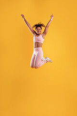 A young woman in a pink athletic outfit joyfully jumps with her arms raised, showcasing excitement and energy. The vivid yellow background enhances her dynamic movements during the activity.