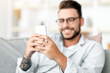 Happy millennial guy enjoying video chat with lover, looking at smartphone screen and smiling, free space. Excited young man holding cellphone while resting on sofa in living room