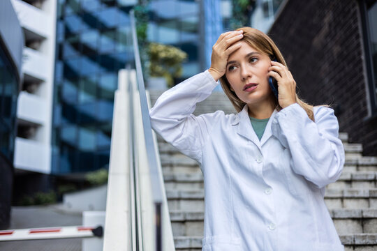 A concerned female healthcare worker in a lab coat using phone, possibly coordinating patient care, against the backdrop of a modern medical facility. Worried doctor making a call outside hospital.