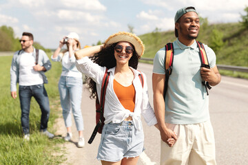 Cool young black guy and his girlfriend traveling together with multinational friends, walking along road, hitchhiking during summer vacation. Autostop journey, tourism and friendship concept