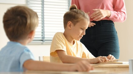 Beautiful teacher explaining test while caucasian boy doing classwork and listening explanation. Cute child looking at teacher while taking a notes or writing answer in paper at classroom. Pedagogy.