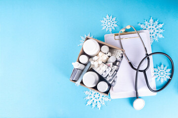 Winter cold and flu viruses therapy and treatment. Box with first aid kit medicines, pills, sprays in female hands, with artificial snowflakes decor and sweater on blue background