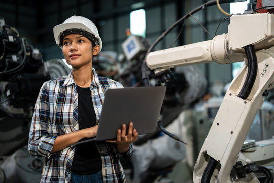 Professional female engineer wearing a safety helmet, holding a laptop to monitor and analyze a robotic arm in an industrial factory, showcasing advanced automation, precision engineering, and modern 