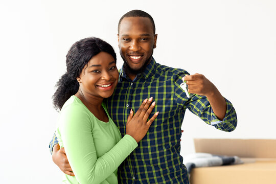 Happy african american spouses demonstrating home keys at camera and embracing, joyful black couple standing among unpacked cardboard boxes, celebrating moving in their new house, copy space