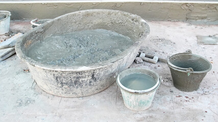 Fototapeta premium Close-up view of cement mixing tools at a construction site, featuring a large basin filled with mixed cement, two smaller buckets and surrounding concrete work surfaces.
