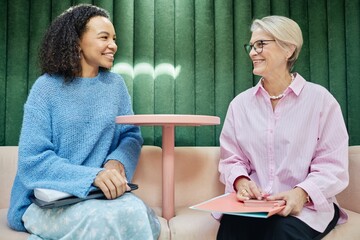 Smiling young multiethnic woman talking to senior female colleague exchanging work experience or...