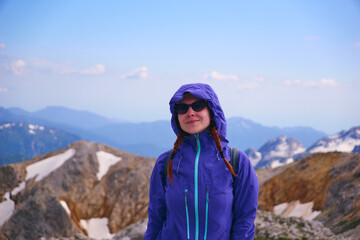 A young girl in sunglasses smiles and looks at the camera against the background of snowy mountain peaks. Mountain trekking and mountaineering in summer