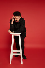 A young handsome man leans on a stool, posing thoughtfully against a bold red backdrop.