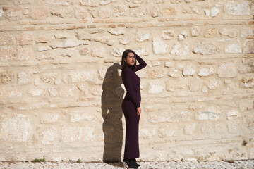 Beautiful young woman dressed in elegant violet dress posing on a stone wall. The woman is serious and has the sun on her face and puts a hand on her head and looks straight ahead.