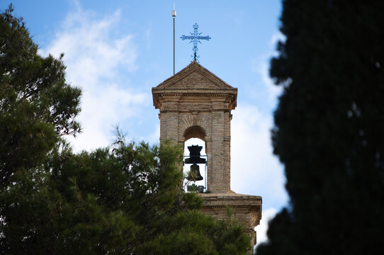 church steeple with bronze bell, on the top a wrought iron cross. In the background the blue sky and white clouds.