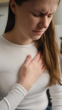 Vertical footage of unhealthy young caucasian woman having severe chest pain while sitting alone on couch, sad worried female suffers from heartache at home, infarction or heart disease concept