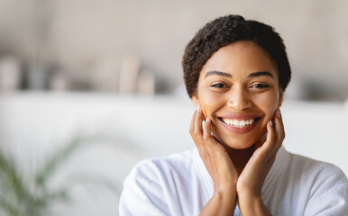 A happy young woman wearing a white spa robe smiles brightly while touching her face. She is inside a peaceful wellness center, enjoying a moment of relaxation and self-care in a calm atmosphere.