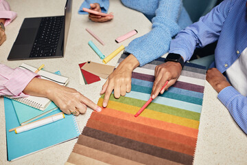 Cropped shot of diverse team of designers choosing fabric among various color samples on tabletop while working collaboratively on new project at workplace, copy spac
