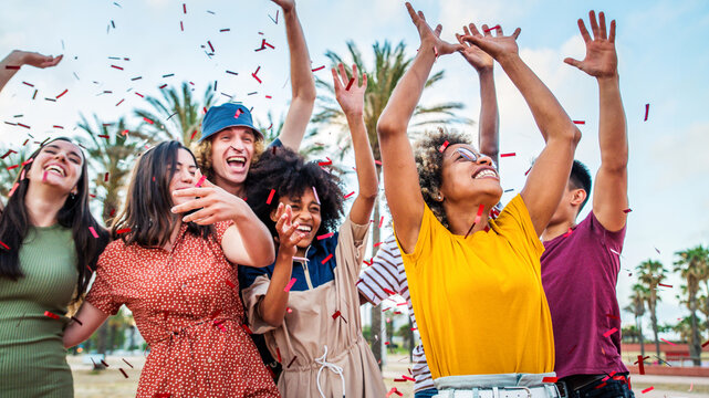 Group of friends enjoying party throwing confetti in the air - Multicultural young students having fun celebrating and laughing out loud outdoors - Youth, friendship and summertime concept