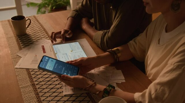 Side cropped shot of anonymous spouses paying taxes and overdue bills online sitting at table, husband using tablet computer and wife holding smartphone, solving financial problems