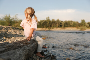Cute little girl sitting on tree trunk near river. Child enjoying beautiful nature