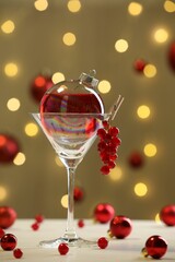 Obraz premium Creative presentation of Christmas cocktail in bauble and glass on white table against dark beige background with blurred lights