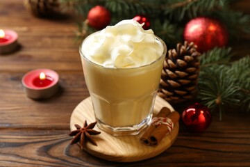 Tasty Christmas cocktail (eggnog) in glass, festive decor and burning candles on wooden table, closeup
