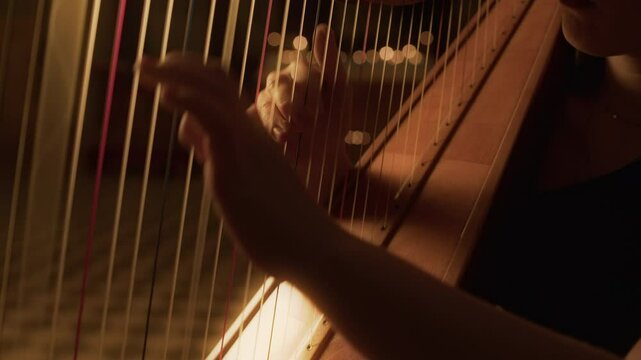 Cropped side view shot of young woman playing masterfully classical harp illuminated by soft warm glow in charming dim light of candles in blurred background