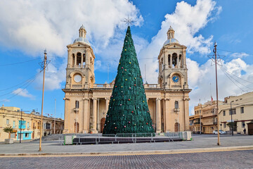 Fototapeta premium Christmas tree stands in front of historic Maltese church with twin bell towers, decorated for festive season under bright blue sky