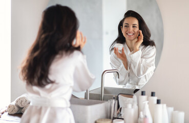 A young brunette woman smiles at her reflection while applying moisturizer in her stylish bathroom. She is dressed in a cozy robe, surrounded by various skin care products on the counter.