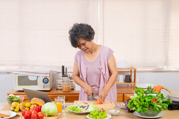 Senior Asian woman with short gray hair preparing healthy food in kitchen, using laptop listen to music or salad recipe research, modern lifestyle silver hair female in 70s friendly with technology