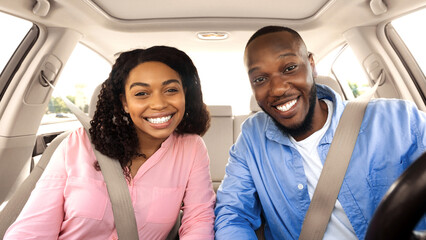 Front portrait of smiling beautiful African American couple sitting in modern luxury car, happy excited young man and woman posing looking at camera, enjoying road trip or buying new car