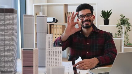 Young Indian male architect in office designing modern city architecture, gesturing okay symbol confidently at camera. Focus on creativity and innovation. Architectural models and laptop visible.
