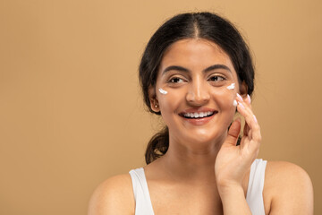 A young Indian woman applies skincare cream to her face while smiling in a bright and calming indoor setting. Her focus is on skincare and self-pampering, showcasing her beauty routine, copy space