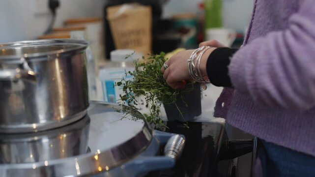 A woman is cooking in a kitchen with a pot on the stove. She is wearing a purple sweater and has a plant in her hand