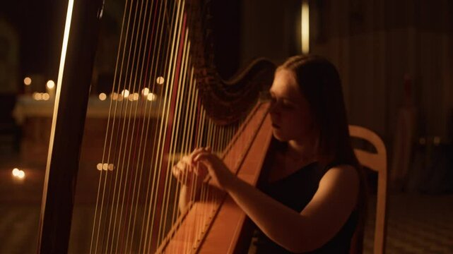 Zoom-in with slowmo shot of shadowed young Caucasian woman in black dress playing classical harp with passion and confidence sitting on chair illuminated by soft charming glow of lit candles of church