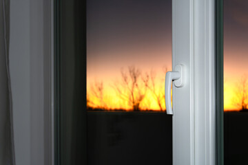 A Warm Evening at Home: A closed window with a modern plastic handle and a view of the sunset. Light shadows from tree branches against the bright sky add depth to the image.