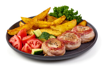 Grilled bacon-wrapped pork medallions with potato wedges, carrot salad, and fresh vegetables on a black plate, isolated on white background.