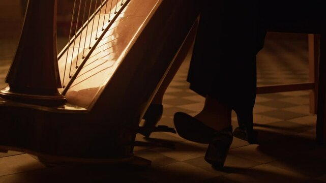 Ground level shot of female harpist playing classical harp illuminated by soft dim light and pressing harp pedals with her feet staying in shadow