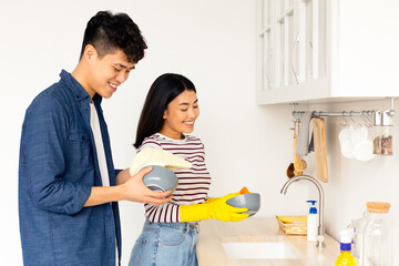 Happy asian guy helping his cheerful girlfriend with washing dishes, side view, panorama with copy...