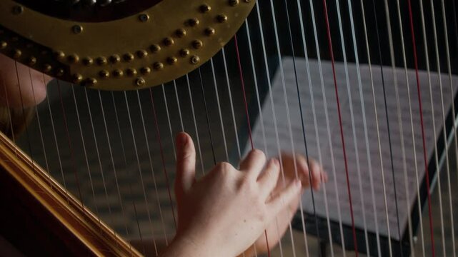 Slowmo cropped shot of young female musician practicing harp playing in dark room with music stand with score in blurred background